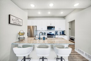 A modern kitchen with white cabinets, a granite countertop, stainless steel appliances, and a bar area with white stools.