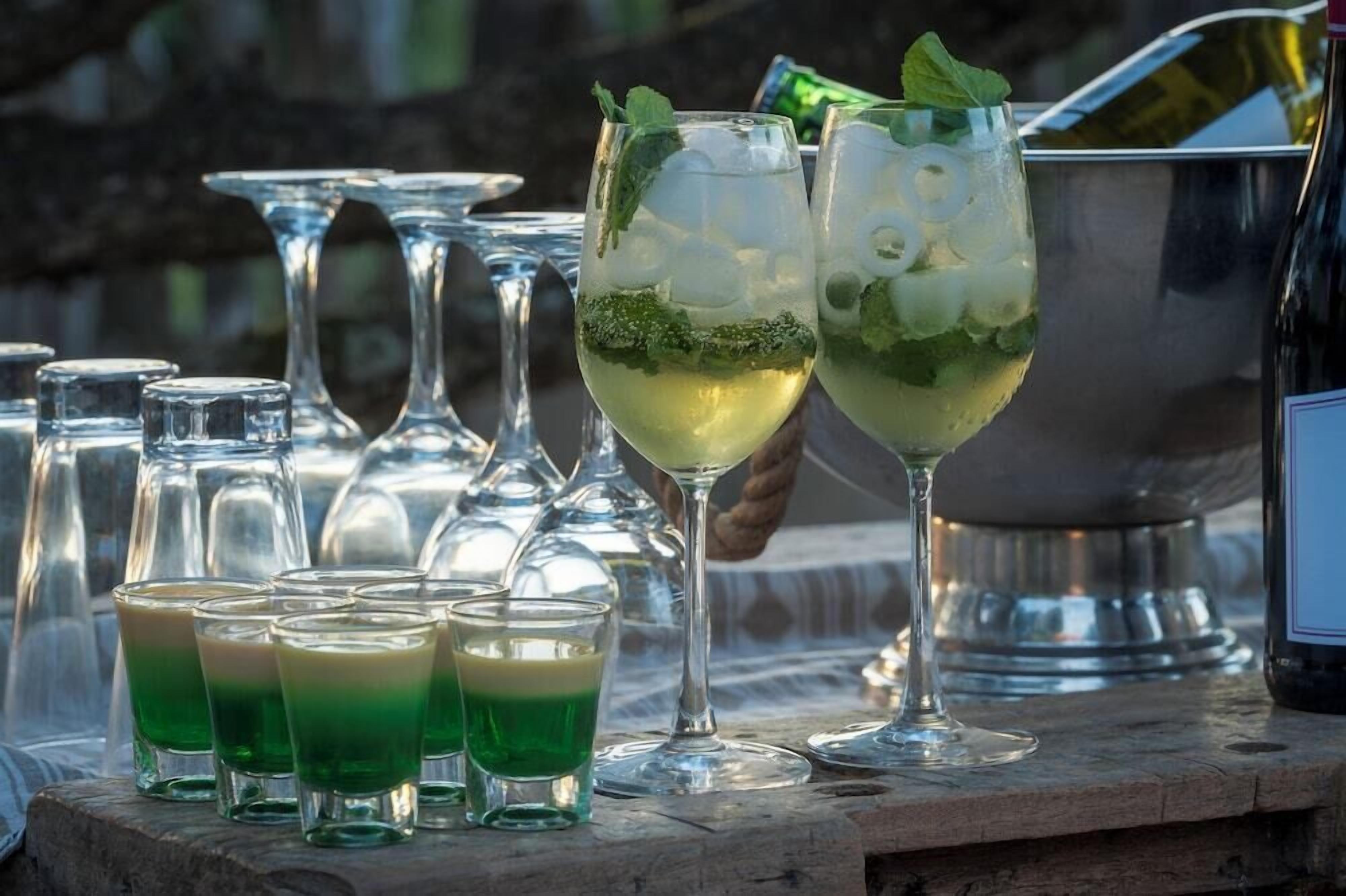 Un bar con vasos de una bebida verde, una botella y un recipiente metálico.