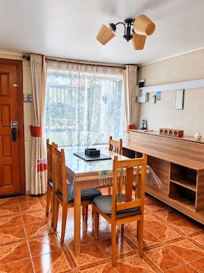 A dining area with a wooden table and chairs, a ceiling light fixture, and a view through a window with curtains.