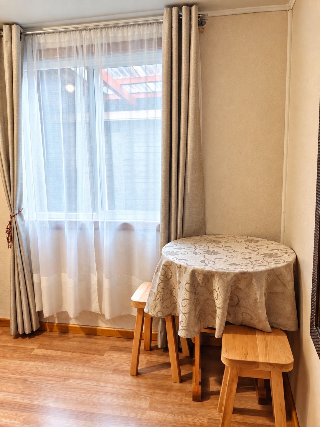 A room with a round table and bench, a window with sheer curtains, and a view of a building outside.