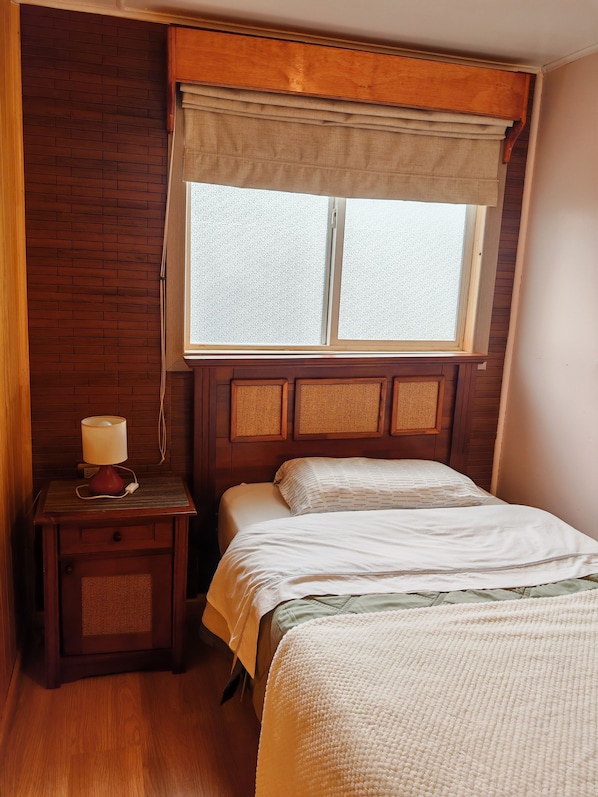 A bedroom with a wooden headboard, a bedside table with a lamp, a window with a view, and a bed with a white comforter.