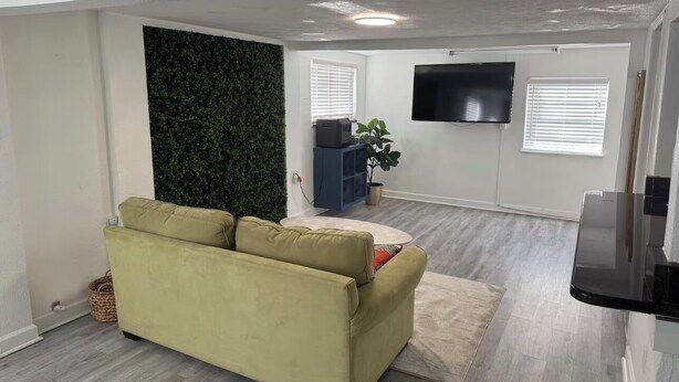 Una sala de estar con sofá, televisor de pantalla plana, planta y ventana con persianas.