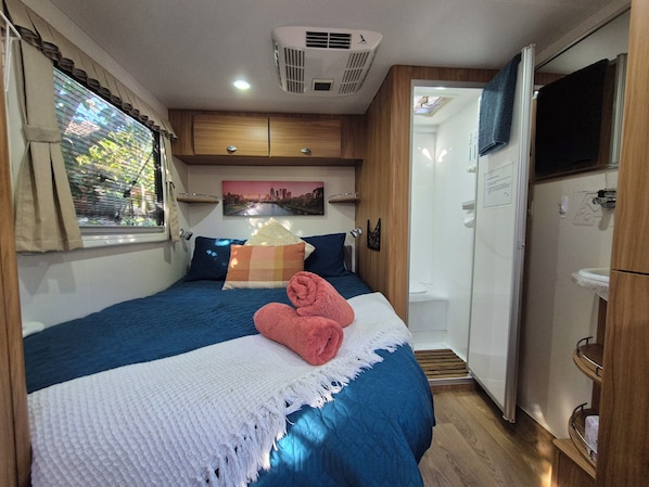 A bedroom with a bed, a window with curtains, a wall-mounted air conditioner, a painting, and a bathroom with a sink and mirror.