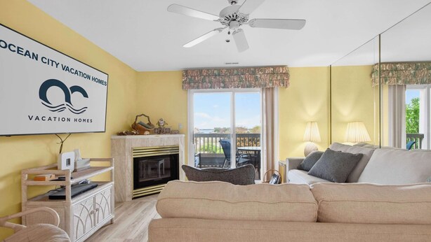 A living room with a sofa, a fireplace, a ceiling fan, and a view of the outdoors. - Bay Watch 1 203A, Outdoor Pool, Beautiful Condo (Ocean City)