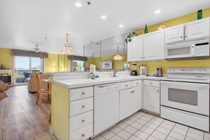 A modern kitchen with white cabinets, a microwave, and a stove. There is a dining area with a chair and a table. - Bay Watch 1 203A, Outdoor Pool, Beautiful Condo (Ocean City)