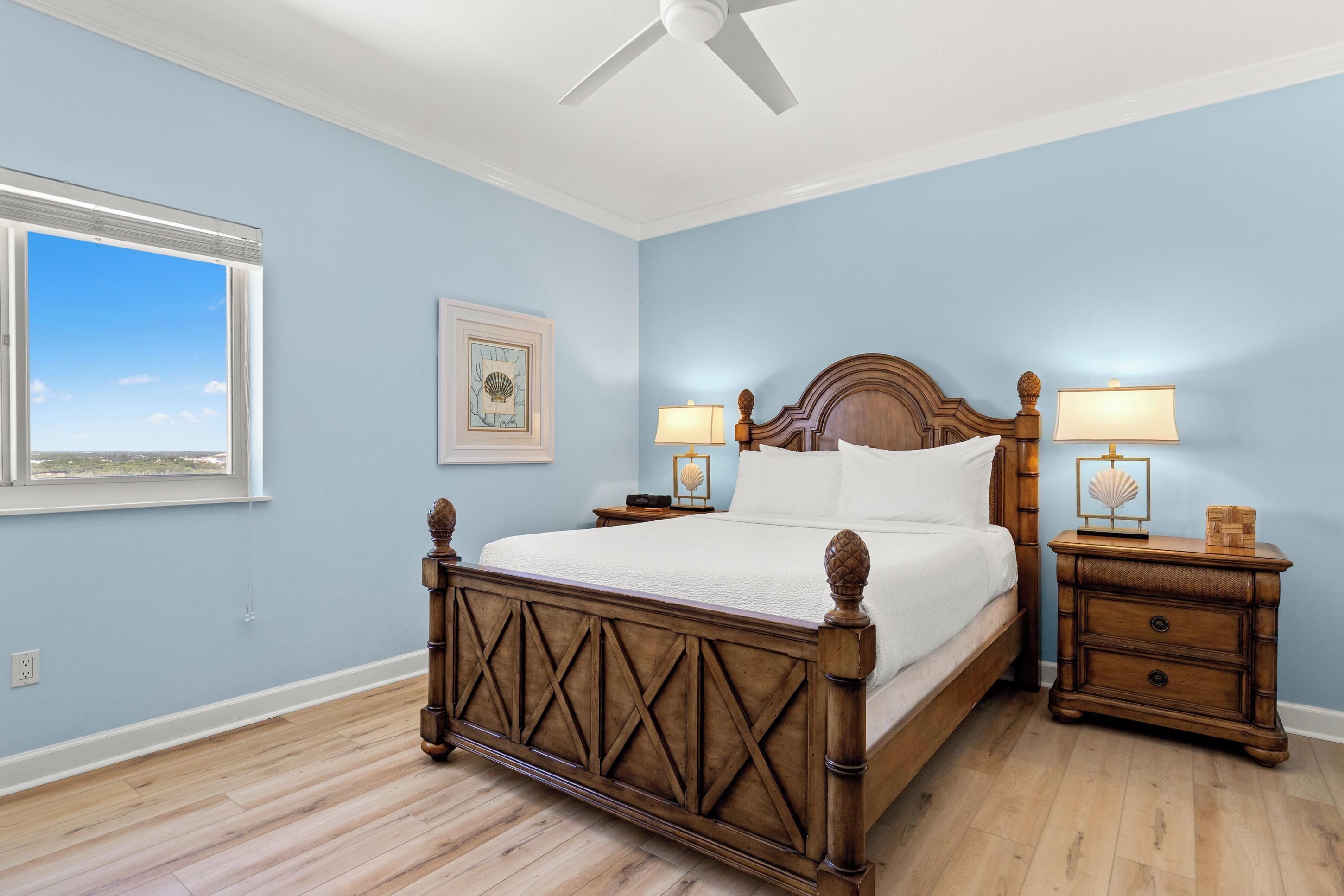 A bedroom with a wooden bed, two bedside tables with lamps, a ceiling fan, a window with blinds, and a framed picture on the wall.