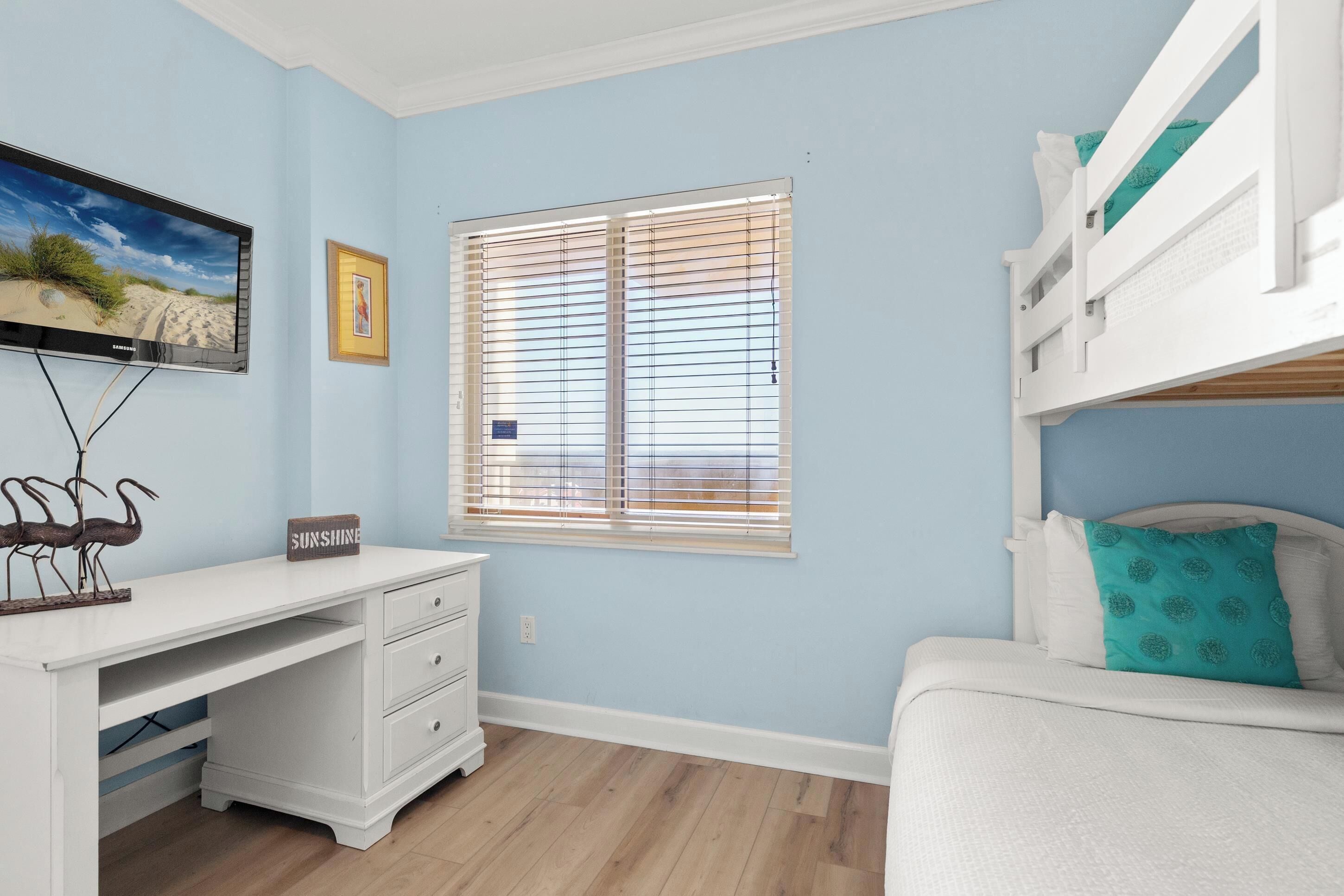 A bedroom with a bunk bed, a desk with drawers, a television, and a window with blinds.