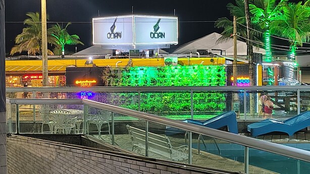 A nighttime view of a waterfront area with illuminated billboards, palm trees, and a brick building facade. - Beachfront apartment in Praia Grande, right on the sand. (Praia Grande)