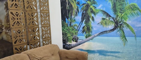 Un sofá color arena en una habitación con un gran mural tropical, un separador decorativo de madera y piso de baldosas.