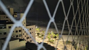 A view through a chain-link fence of a building with multiple floors, a street with palm trees, and a beach in the background. - Beachfront apartment in Praia Grande, right on the sand. (Praia Grande)