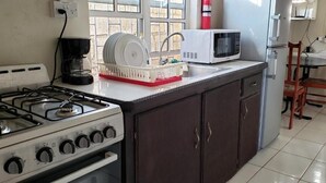 A kitchen with a gas stove, a microwave, a dish rack, and a dining area with chairs and a table. - Roseau Beach Hostel  (Roseau)