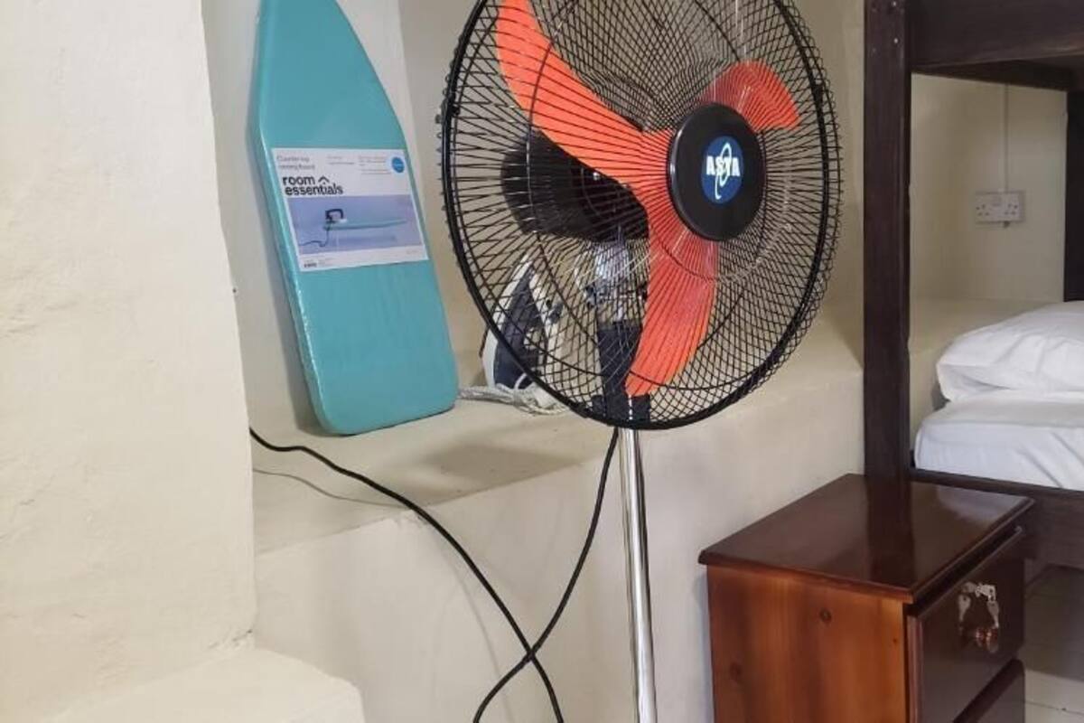 Un llit superposat amb un capçal de fusta, una petita taula de nit de fusta i un ventilador de peu amb disseny vermell i negre.