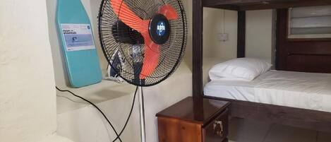 Una cama litera con cabecera de madera, una mesita de noche de madera pequeña y un ventilador de pie con diseño rojo y negro.