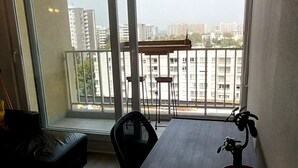 A room with a dining table, chairs, a potted plant, and a view of buildings through large windows.