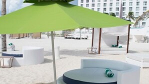 Ein Sitzbereich am Strand mit einer runden weißen Bank, einem grünen Sonnenschirm und einer weißen Markise im Hintergrund.