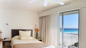 Ein Hotelzimmer mit einem Bett, einem Nachttisch, einer Vase mit Blumen, einem Fenster mit Blick auf den Strand und einem Deckenventilator.