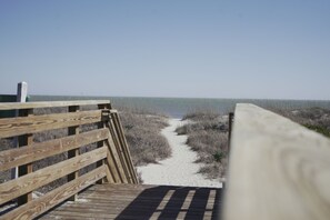 Ein hölzerner Steg führt zu einem Strand mit Sand und vereinzelter Vegetation.