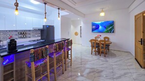 A modern kitchen with a central island, bar stools, a dining area with wooden chairs, a TV mounted on the wall, and a chandelier. - Elite Holiday Homes - Akdital & Aéroport (Fes)