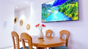 A dining area with a round wooden table, four wooden chairs, a vase with flowers, and a flat-screen TV mounted on the wall displaying a scenic landscape. - Elite Holiday Homes - Akdital & Aéroport (Fes)
