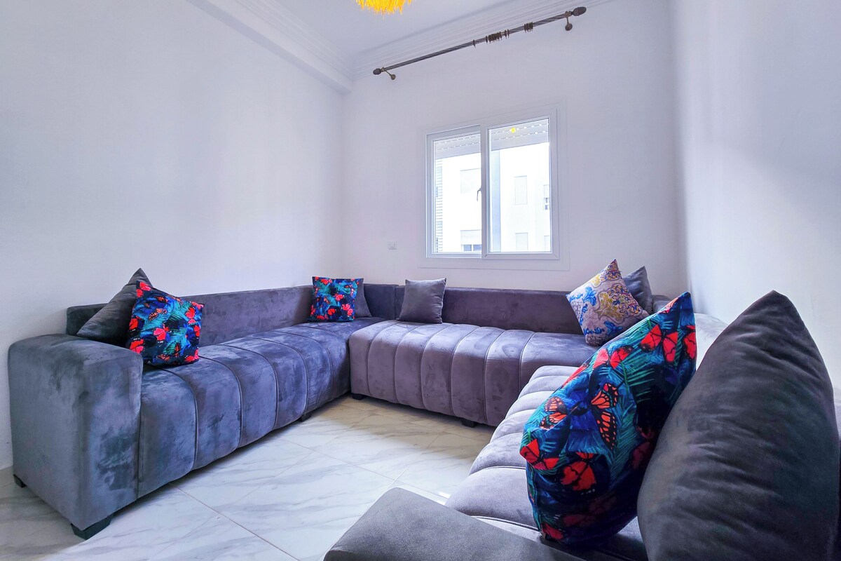 A modern living room with a grey sectional sofa, colorful pillows, a hanging light fixture, and a window with blinds.