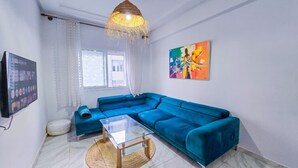 A living room with a sectional sofa, a glass coffee table, a woven rug, a television, and a colorful painting on the wall. - Elite Holiday Homes - Akdital & Aéroport (Fes)