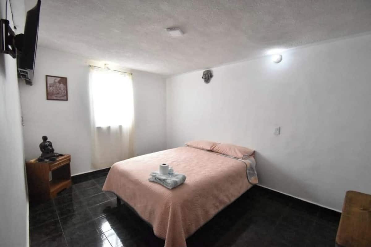 Ein Schlafzimmer mit einem Bett, einem Nachttisch, einem an der Wand montierten Fernseher und einem Fenster mit Vorhängen.