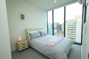 A modern hotel room with a large bed, a bedside table, a nightstand, and a window with a city view. - VTA | Chic 2-BR City Retreat with Ensuite Master (Adelaide)