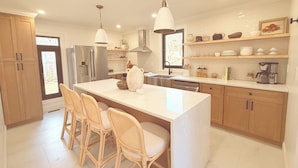 Cocina moderna con una isla central, taburetes de bar, armarios de madera, un refrigerador de acero inoxidable y una cafetera.