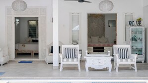 Exterior blanco con ventilador de techo, dos lámparas colgantes de mimbre, una silla blanca con cojines a rayas, una mesita y un aparador blanco con libros y adornos.