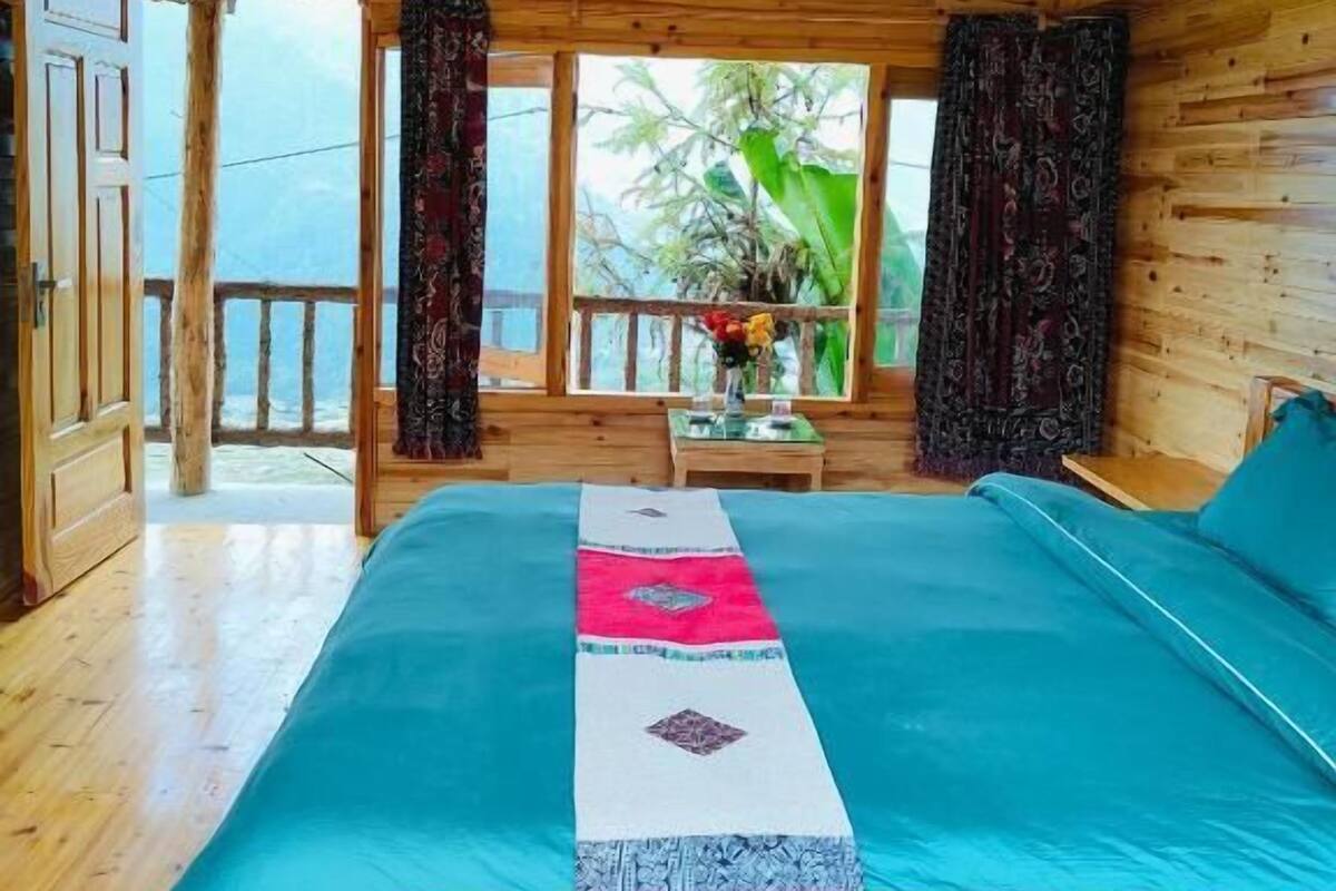 A wooden cabin bedroom with a bed, curtains, a window with a view, and a ceiling light.