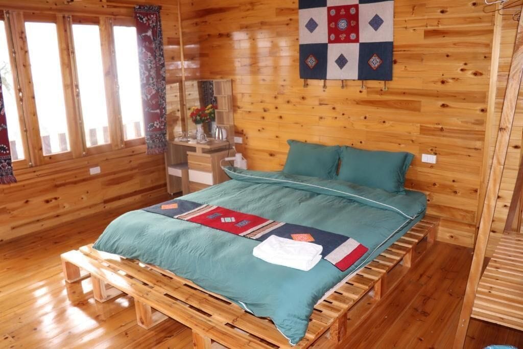 Une pièce de cabane en bois comprenant un lit, une couverture, une tapisserie murale matelassée et une fenêtre avec des rideaux.