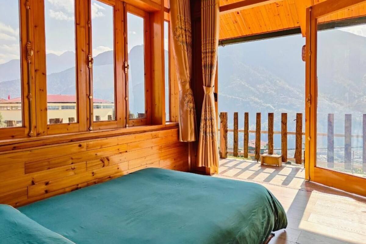 A wooden cabin room with a bed, large windows, and a balcony with a view of mountains.