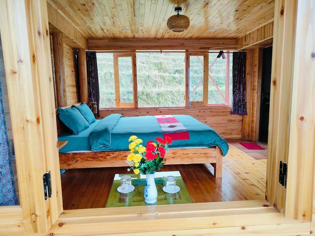 Une chambre dans une cabane en bois, avec un lit, un vase de fleurs et une fenêtre avec des rideaux.