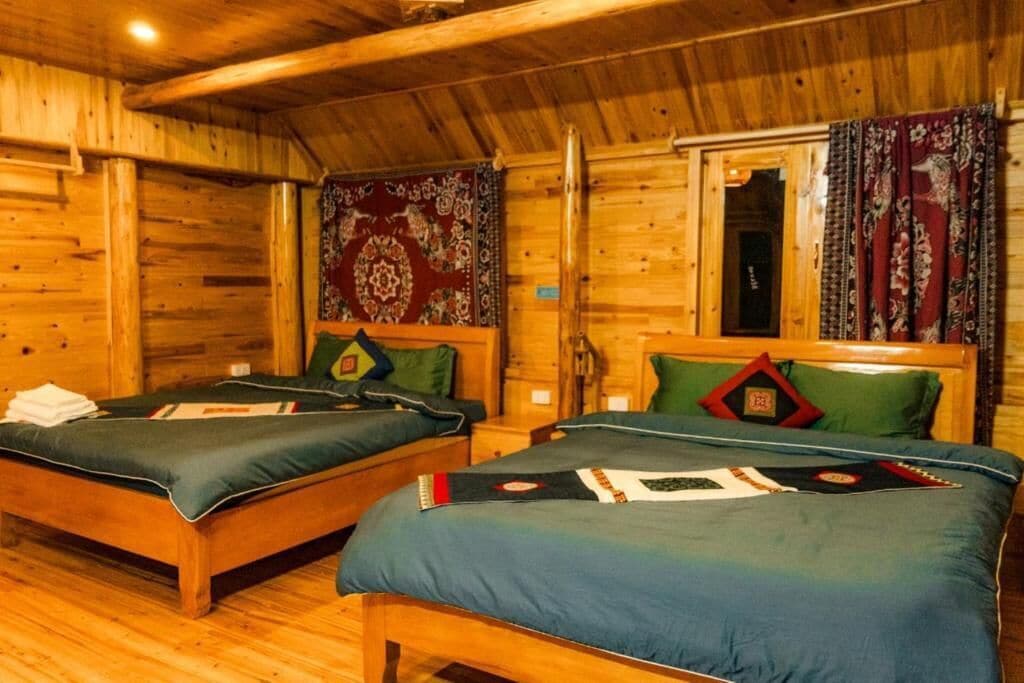 A wooden cabin with two beds, each with a green bedspread and pillows, a wooden ceiling, and a window with curtains.