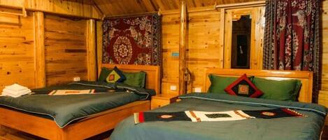 A wooden cabin with two beds, each with a green bedspread and pillows, a wooden ceiling, and a window with curtains.