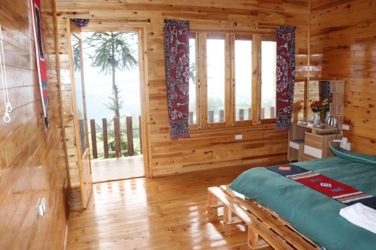 A wooden cabin room with a bed, a bench, a desk, and a view of the outdoors.
