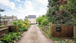 Une allée de gravier mène à une maison dotée d’un portail en bois et d’une boîte aux lettres située sur le côté droit.
