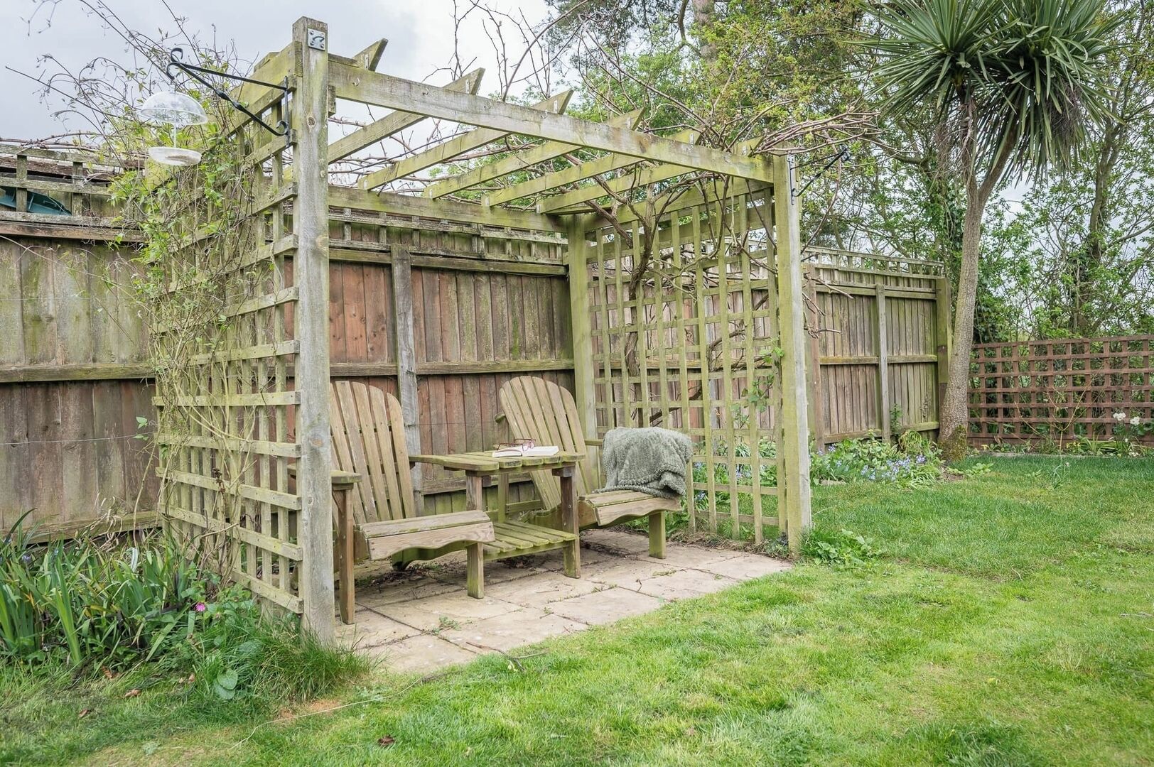 Una pergola in legno con una panchina e un tavolo, circondata da un recinto in legno e vegetazione.