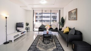Een moderne woonkamer met een flatscreen televisie, een bijzettafeltje met een vaas, een zwarte bank met kussens, een bankje en een groot raam met uitzicht op gebouwen.