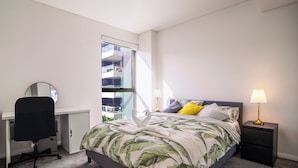 Een slaapkamer met een bed, bureau, stoel, nachtkastje, lamp en een raam met uitzicht op een gebouw.