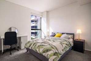 A bedroom with a bed, a desk, a chair, a nightstand, a lamp, and a window with a view of a building. - Spacious 3BR, 10 mins to CBD, 5 mins to Airport (Mascot)