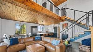 A spacious living area with a large brown leather sectional sofa, a dining table with chairs, a kitchen area, and a staircase with a wooden finish. - Gray Ghost | Hot Tub | Views | Pet Friendly (Rangeley)