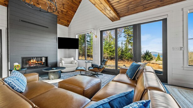 A modern living room with a fireplace, leather sofa, and large windows offering a view of the outdoors. - Gray Ghost | Hot Tub | Views | Pet Friendly (Rangeley)