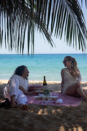 Zwei Personen sitzen am Strand mit einem Picknick, darunter eine Flasche und Gläser.