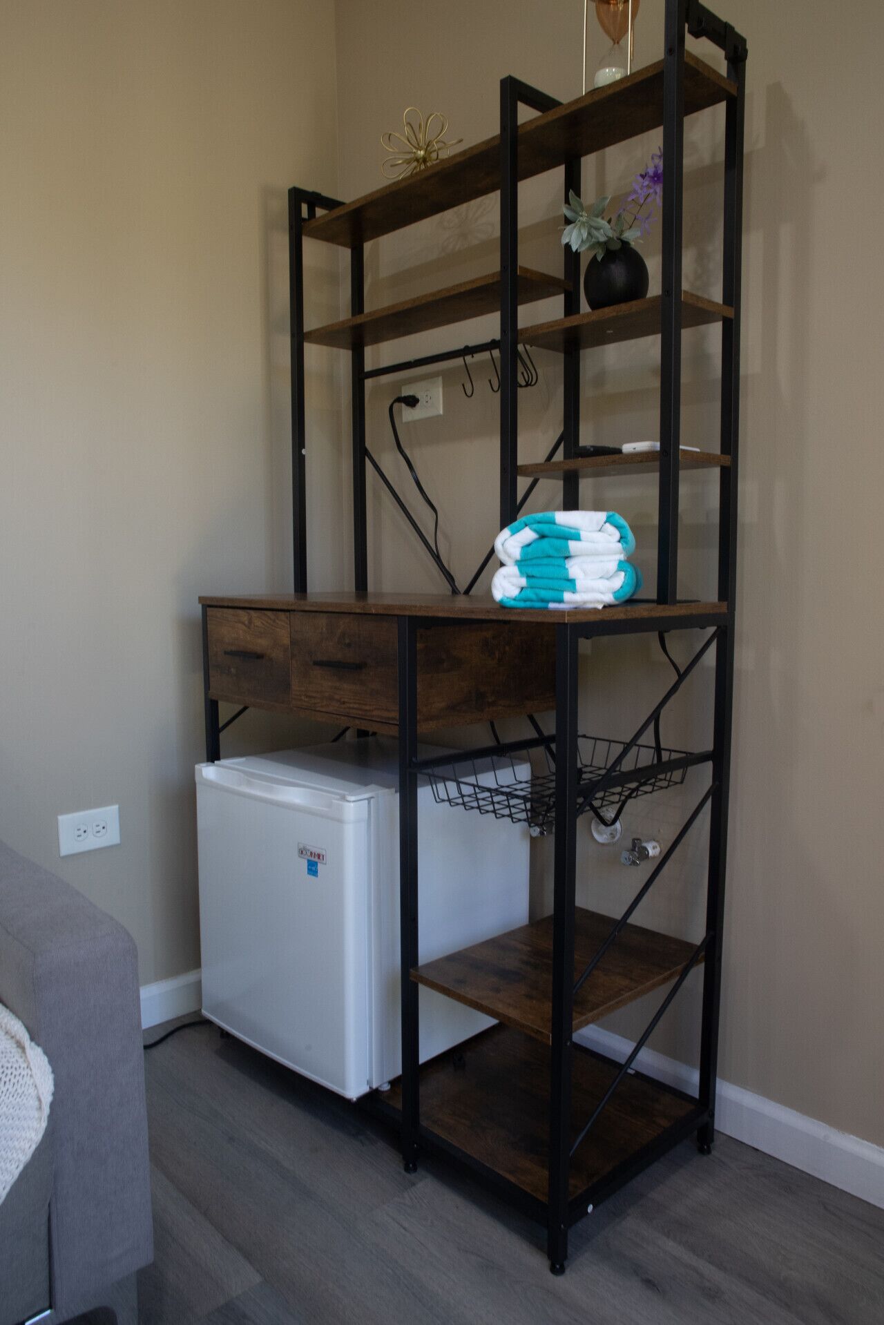 A compact kitchenette with a white mini-fridge, a wooden shelf unit with a towel rack, and a small plant.