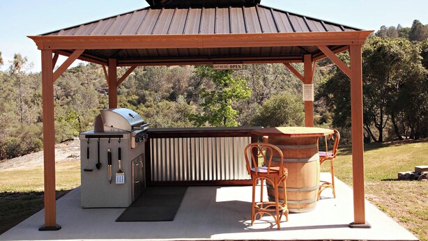 Un espace barbecue extérieur abrité, avec une table et des chaises en bois, un gril et un baril.