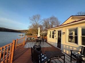 A wooden deck with outdoor seating, a view of the lake, and a building with large windows. - Cozy Cottage on the Lake (Port Jervis)
