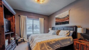 A bedroom with a bed, a television, a window with curtains, a wall tapestry, and a nightstand with a lamp. - McCall Basecamp (Donnelly)