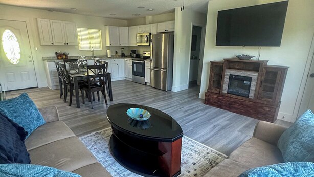 Una sala de estar con sofá, mesa de centro y chimenea. Se observa una cocina al fondo, con armarios y un refrigerador.