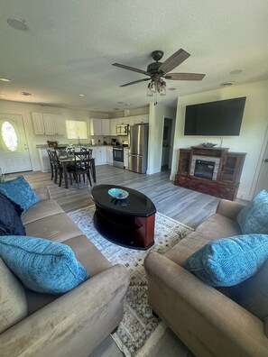 A living room with a sofa, coffee table, and a fireplace. There is a kitchen in the background with cabinets and a refrigerator. - 3BR Home - Prime Pigeon Forge Location - Near Dining & Shops!! (Pigeon Forge)